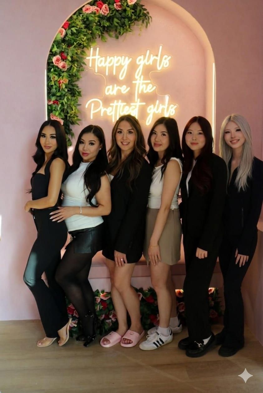 Six women posing before a floral backdrop with a glowing neon sign in a pink room.