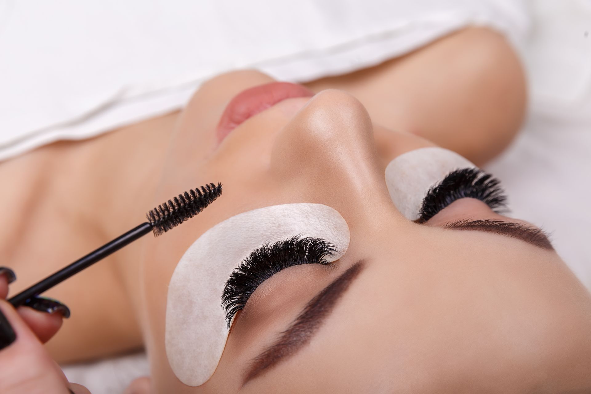 Person lying down with eyelash extensions being applied using a mascara wand and white eye patches