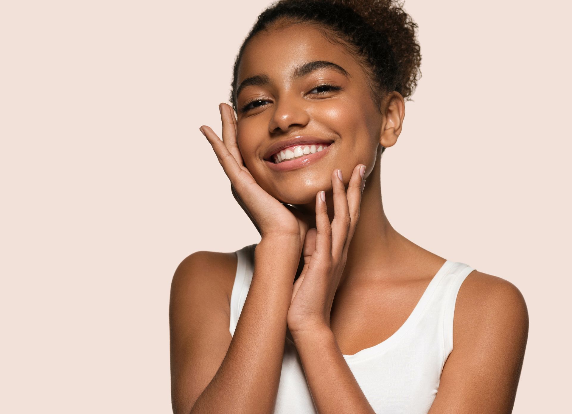Smiling person in a white tank top touching their face against a soft pink background
