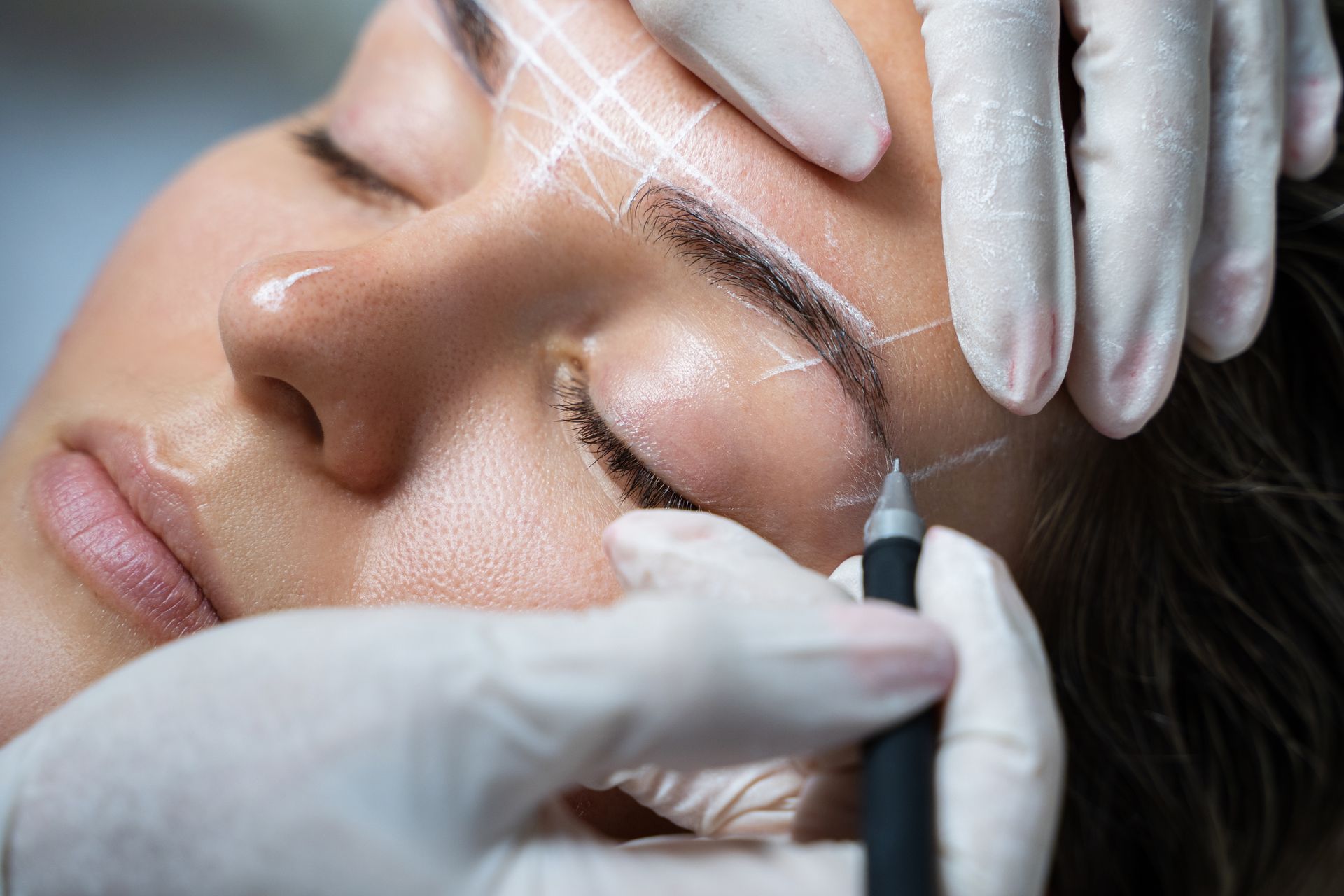 Cosmetic procedure on a client’s eyebrow with a handheld tattoo pen and gloved hands