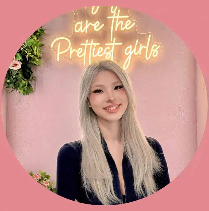 Smiling woman with long blonde hair in a black top against a pink wall with neon sign text.