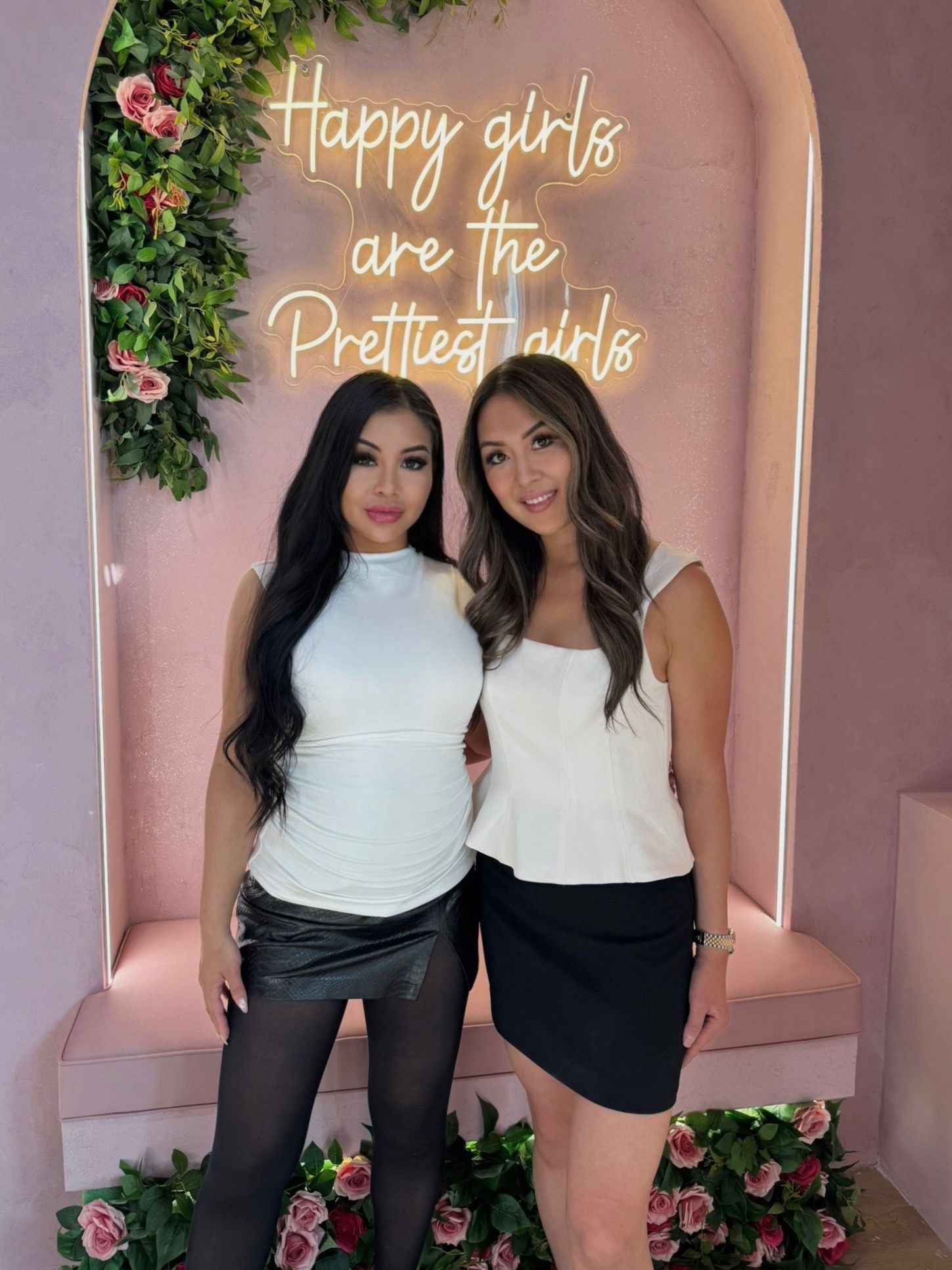 Two women posing in front of a pink floral backdrop with neon sign reading “Happy girls are the prettiest girls”