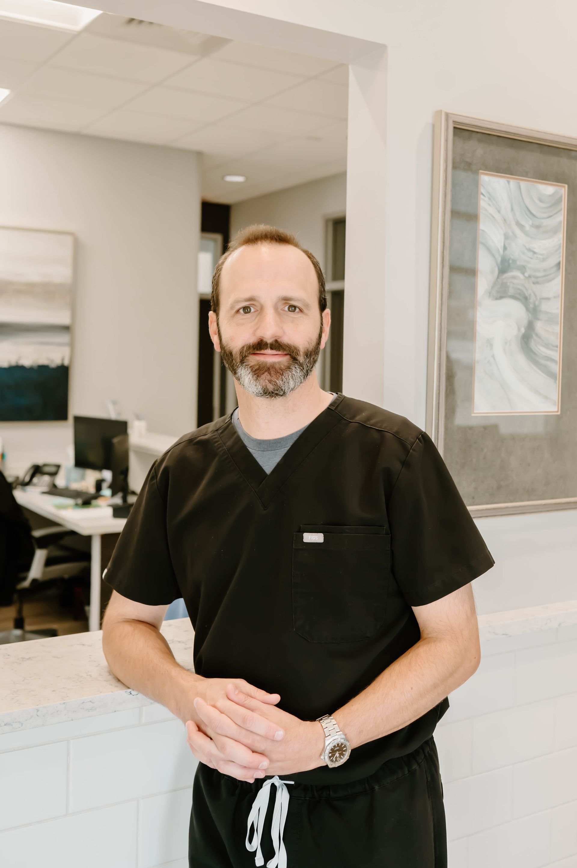 A man with a beard is wearing scrubs and standing next to a counter.