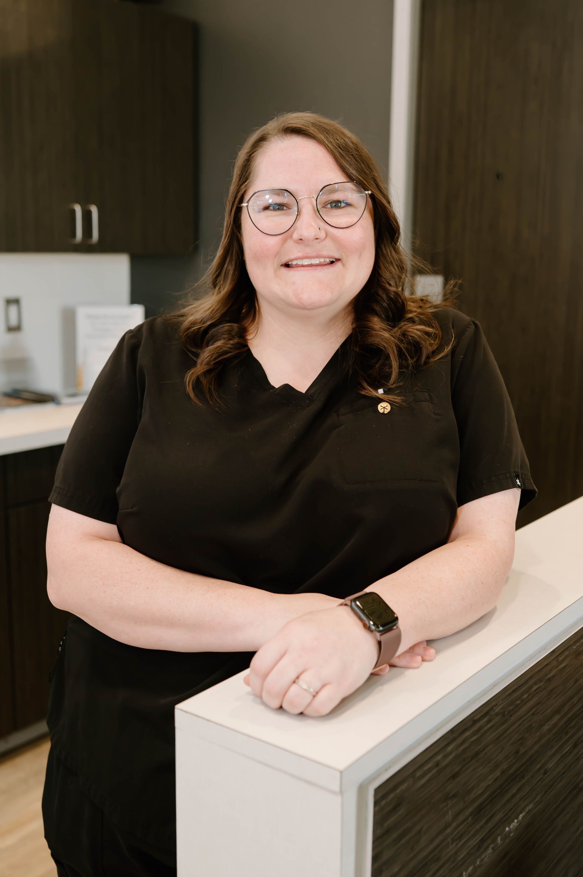 Woman in black scrubs smiles, leaning on a white counter, artwork in background.