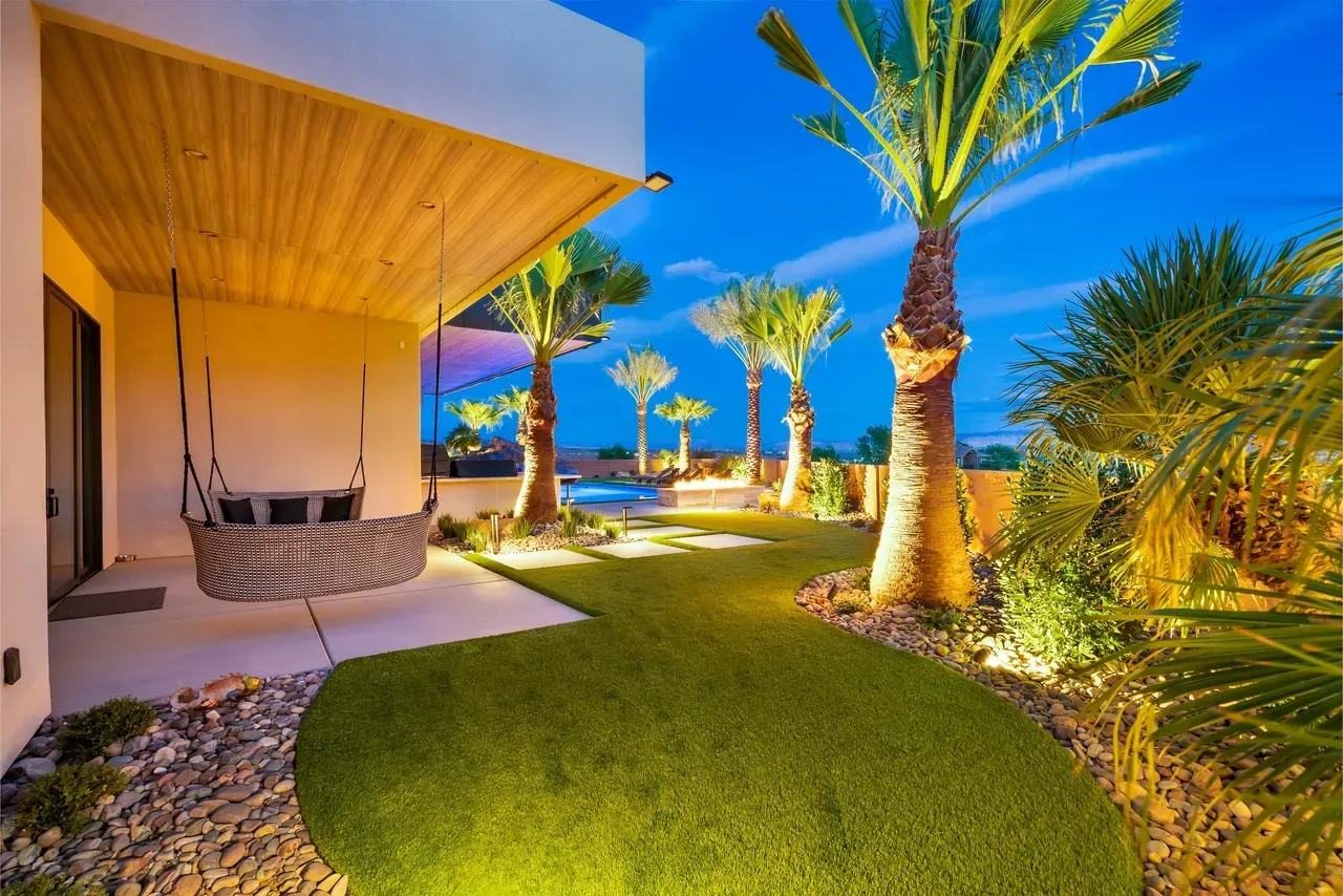 Outdoor patio with a swing, palm trees, and illuminated landscaping at dusk.