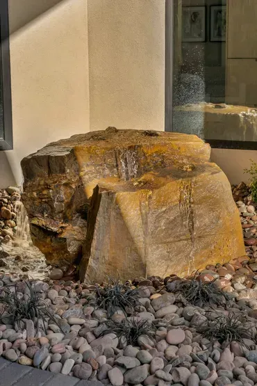 Large, wet, brown rock fountain in a stone garden bed next to a building.