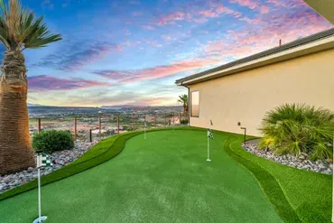 Green putting green with flags, next to a beige house, overlooking a scenic view at sunset.