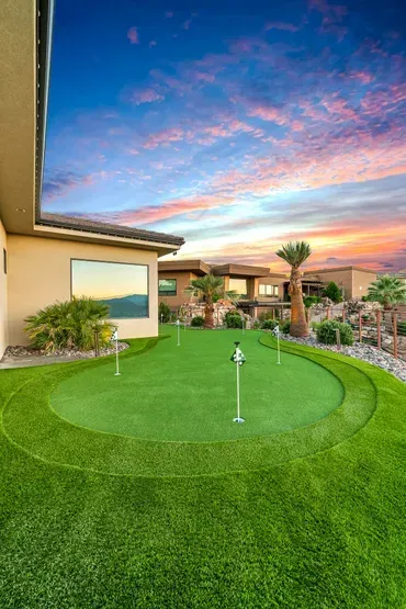 Backyard putting green with flags, surrounded by green grass and desert landscape under a colorful sunset.