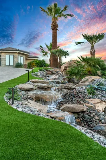 Waterfall feature in a yard with palm trees, green lawn, rocks, and a home at sunset with a colorful sky.