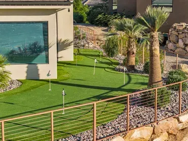 Putting green with flags, next to a modern house with large windows, and palm trees.