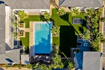 Aerial view of a residential complex with a rectangular pool, green lawns, palm trees, and two buildings.