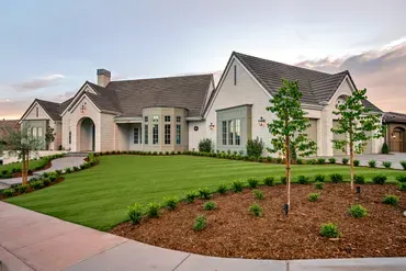 Exterior view of a large, beige house with a manicured lawn and landscaping, under a cloudy sky.