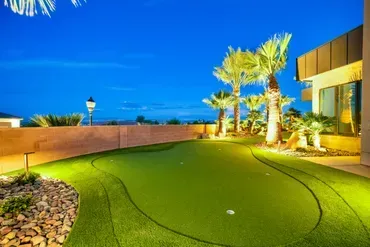 Backyard putting green at night, lit by spotlights. Palm trees and a low wall surround the green.