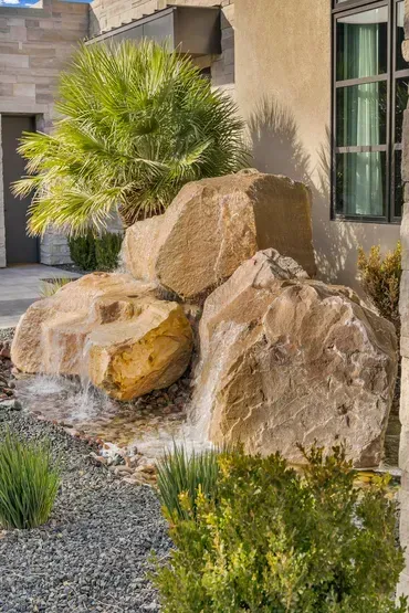 Water cascading over large, tan boulders in a landscaped yard. Green shrubs and a palm tree are nearby.