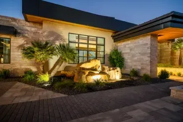 Modern home exterior with illuminated landscaping and rock water feature at dusk.