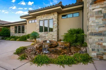 Luxurious house exterior with stone facade, windows, small waterfall, and landscaping under a blue sky.