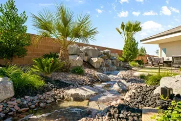 Backyard with a rock waterfall feature, palm trees, and landscaping near a house.
