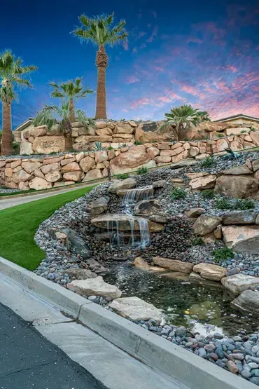 Waterfall cascading down rock formations in front of a palm-tree-lined landscape; sunset sky.