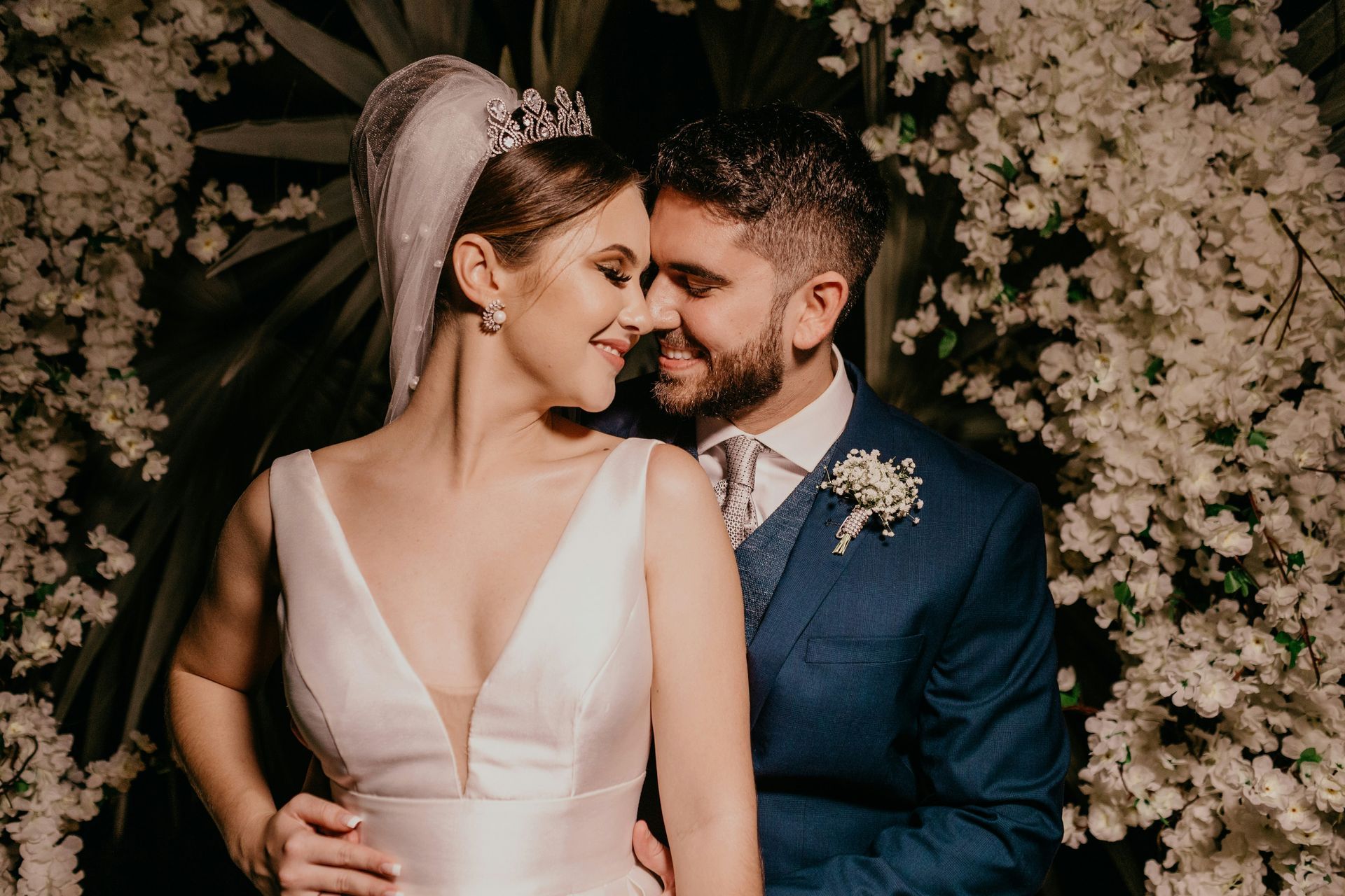 Bride and groom posing closely in front of a floral backdrop, with the bride in white and groom in navy suit