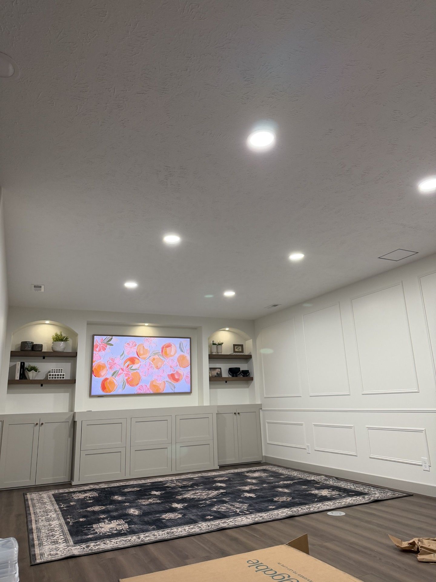 Bright basement media room with a wall-mounted TV, recessed lights, white cabinets, and a patterned rug.