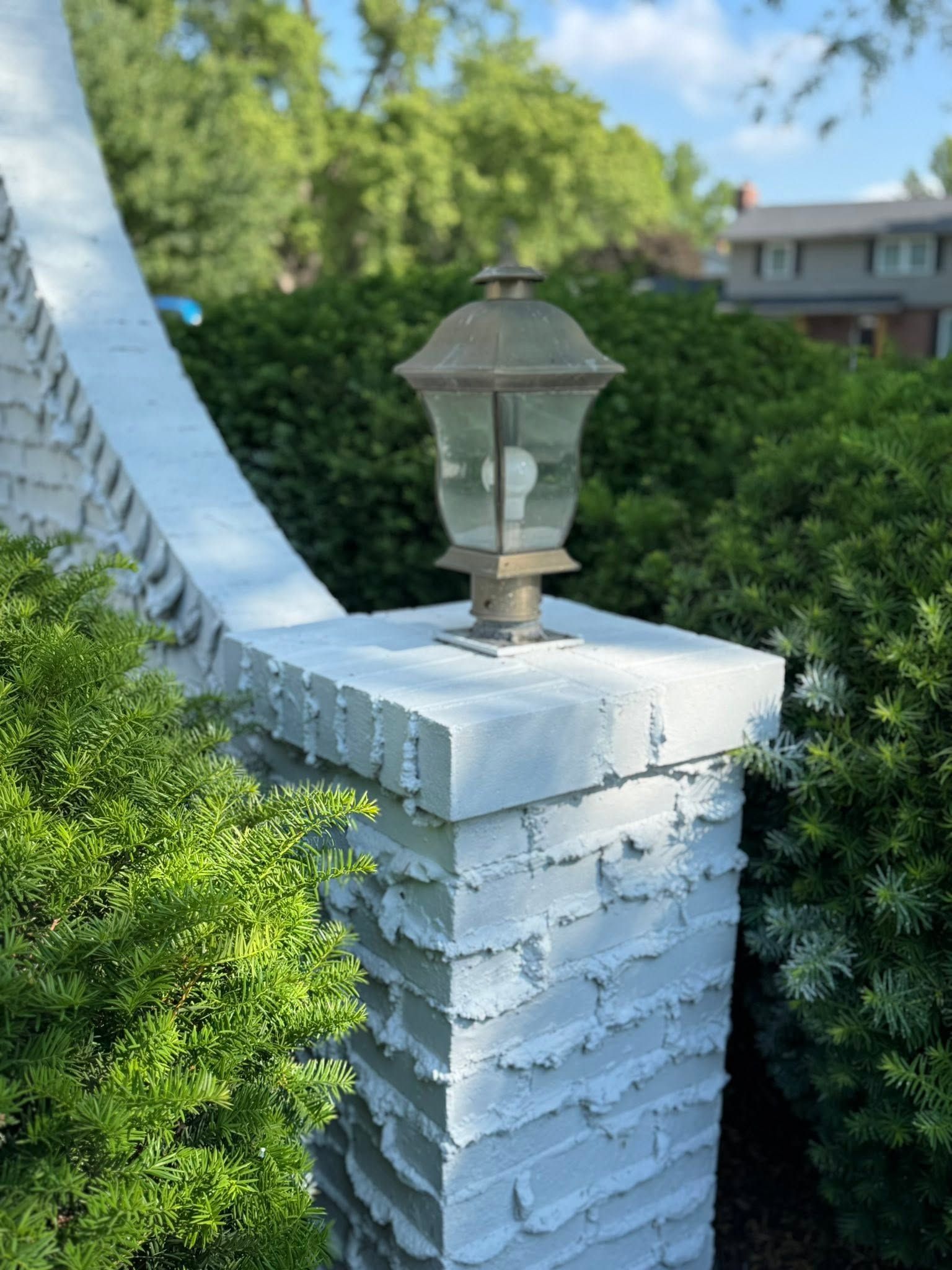 Weathered lantern on a white brick pillar beside a lush green hedge on a sunny day