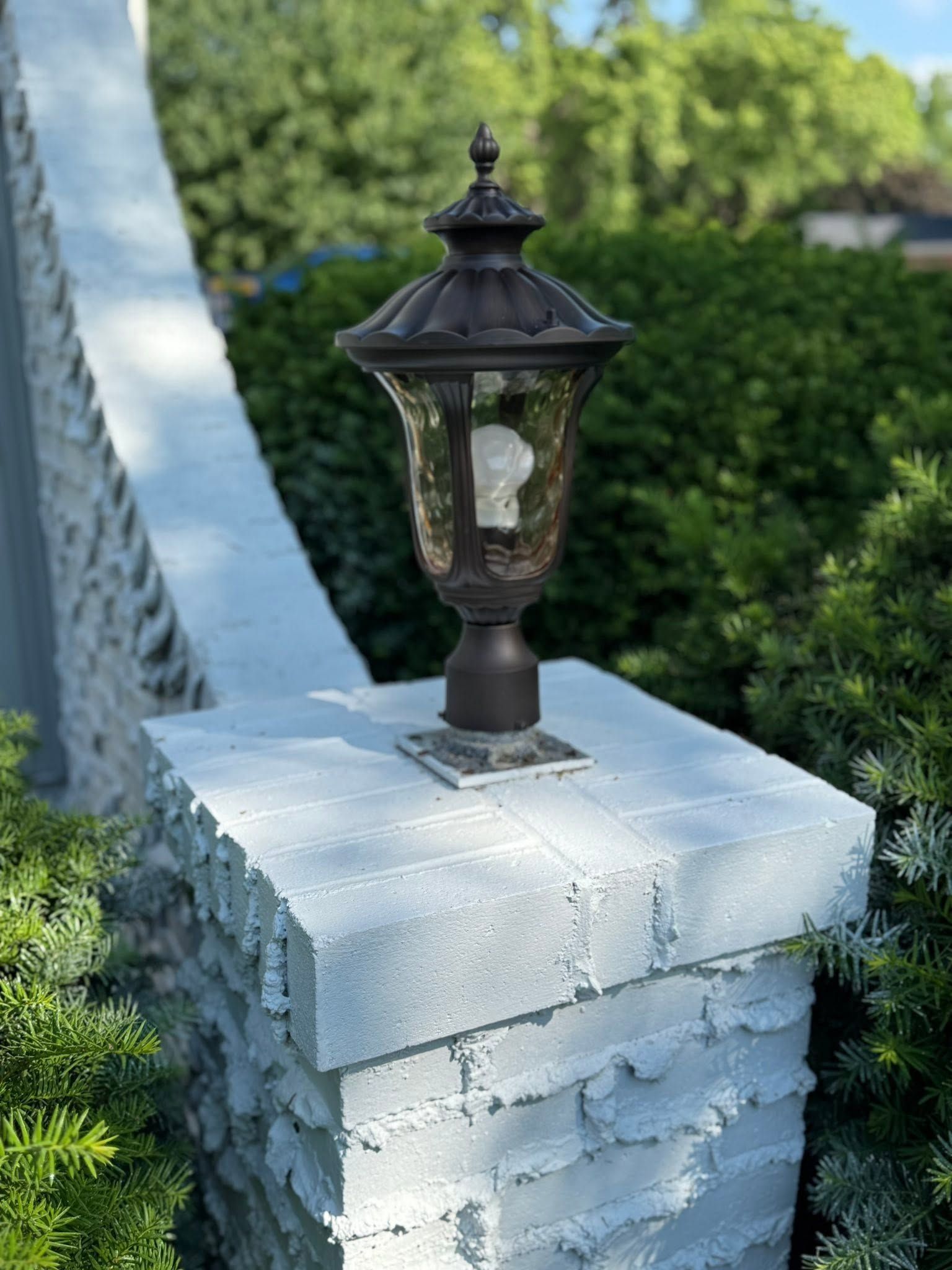 Black lantern-style light on a white brick fence post with green trees in the background