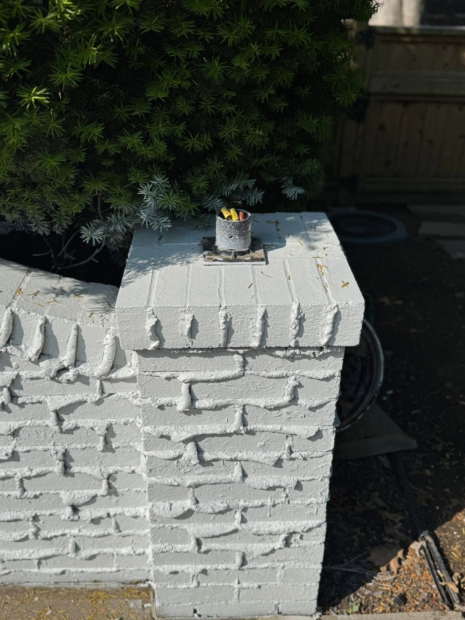 White brick pillar with a small metal can on top, beside a green bush outdoors