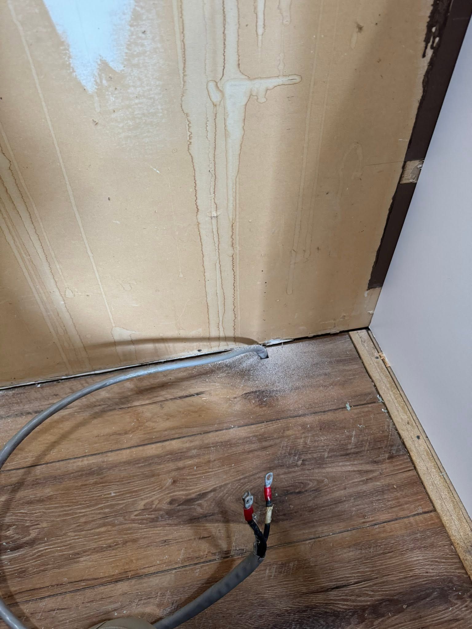 Exposed wires on a wooden floor beside a damaged wall near a baseboard repair area