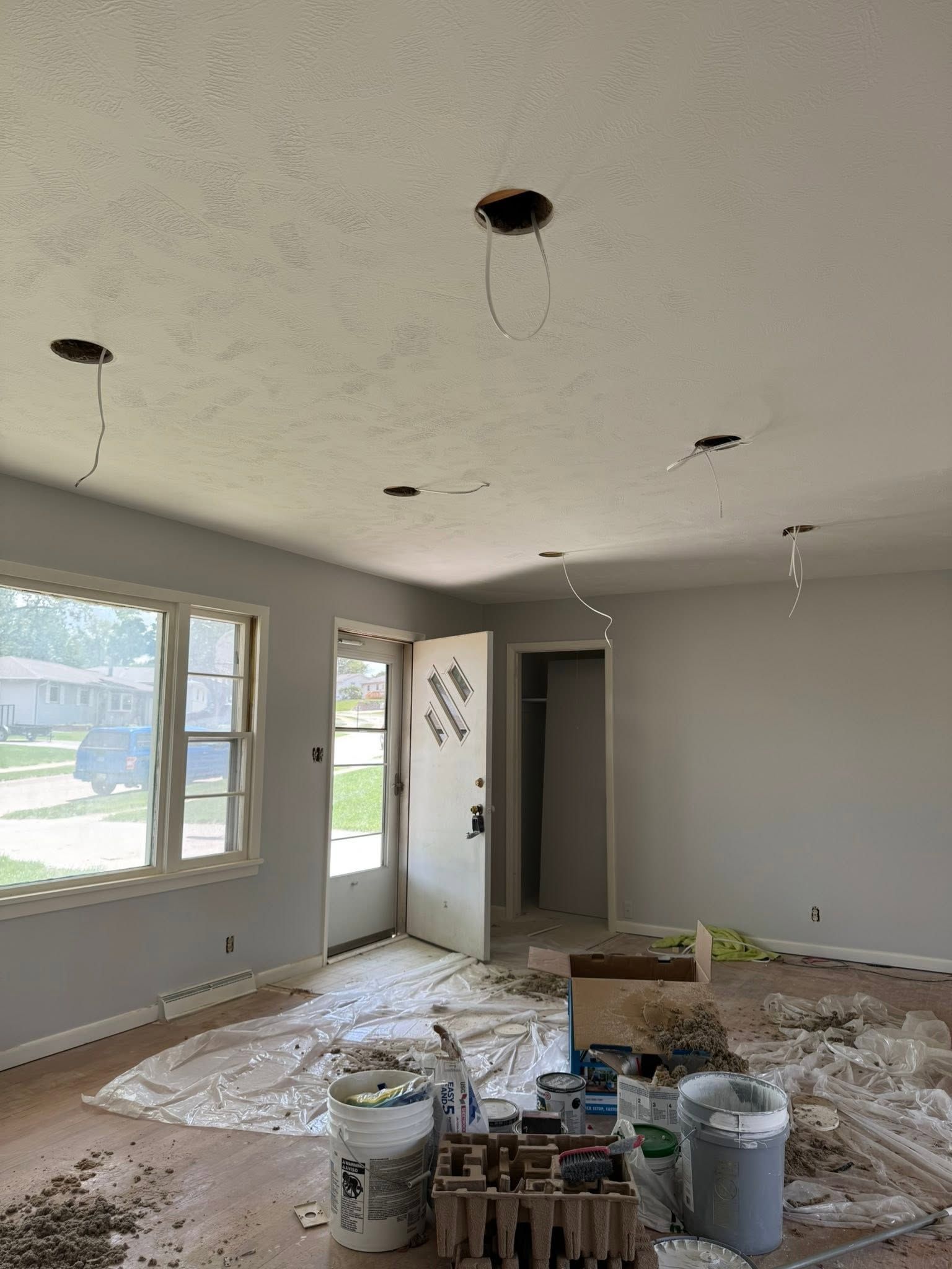 Unfinished living room with drywall, plastic-covered floor, open doorway, and paint cans near windows.