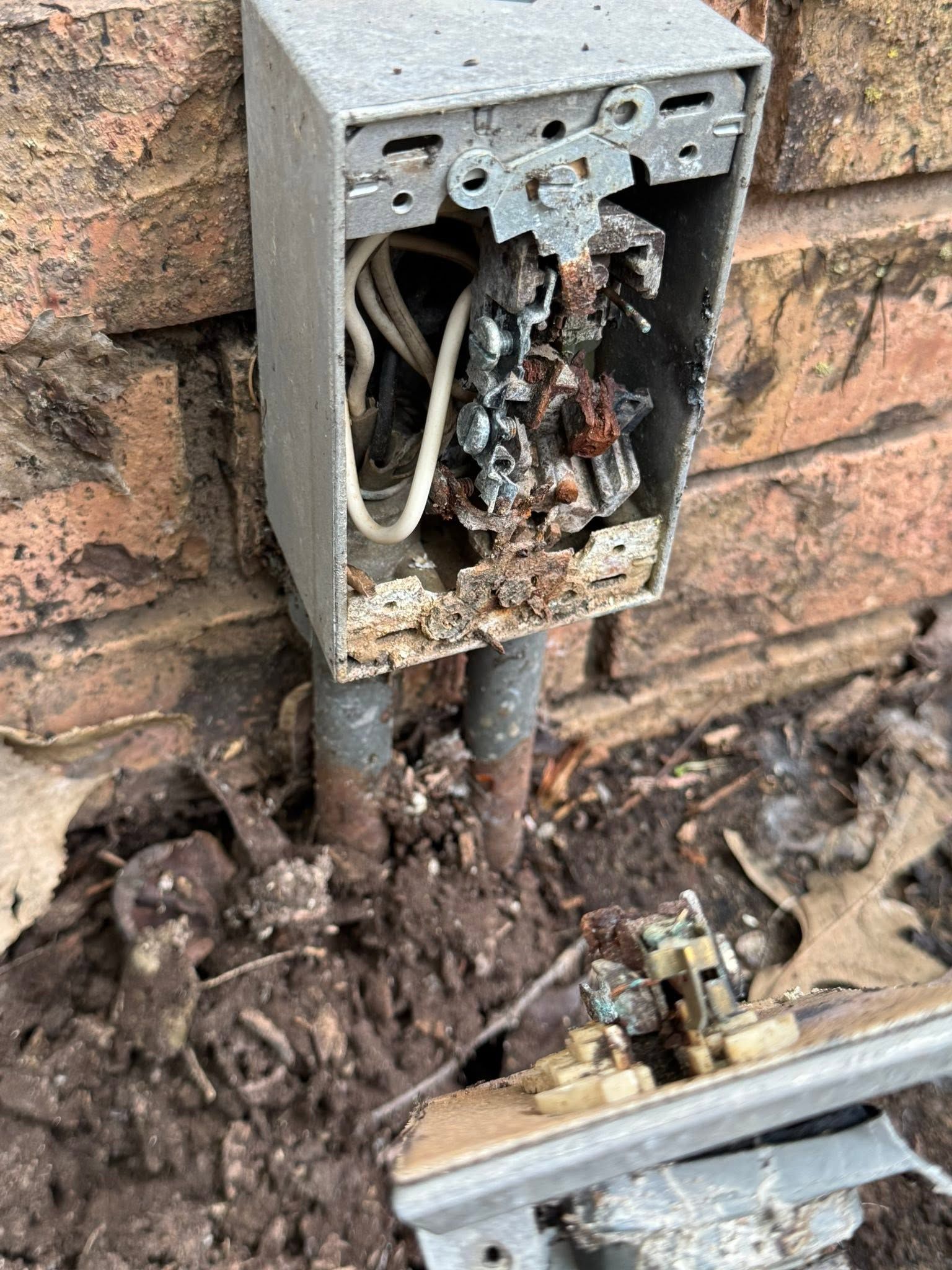 Open outdoor electrical box with exposed wiring and rust against a brick wall