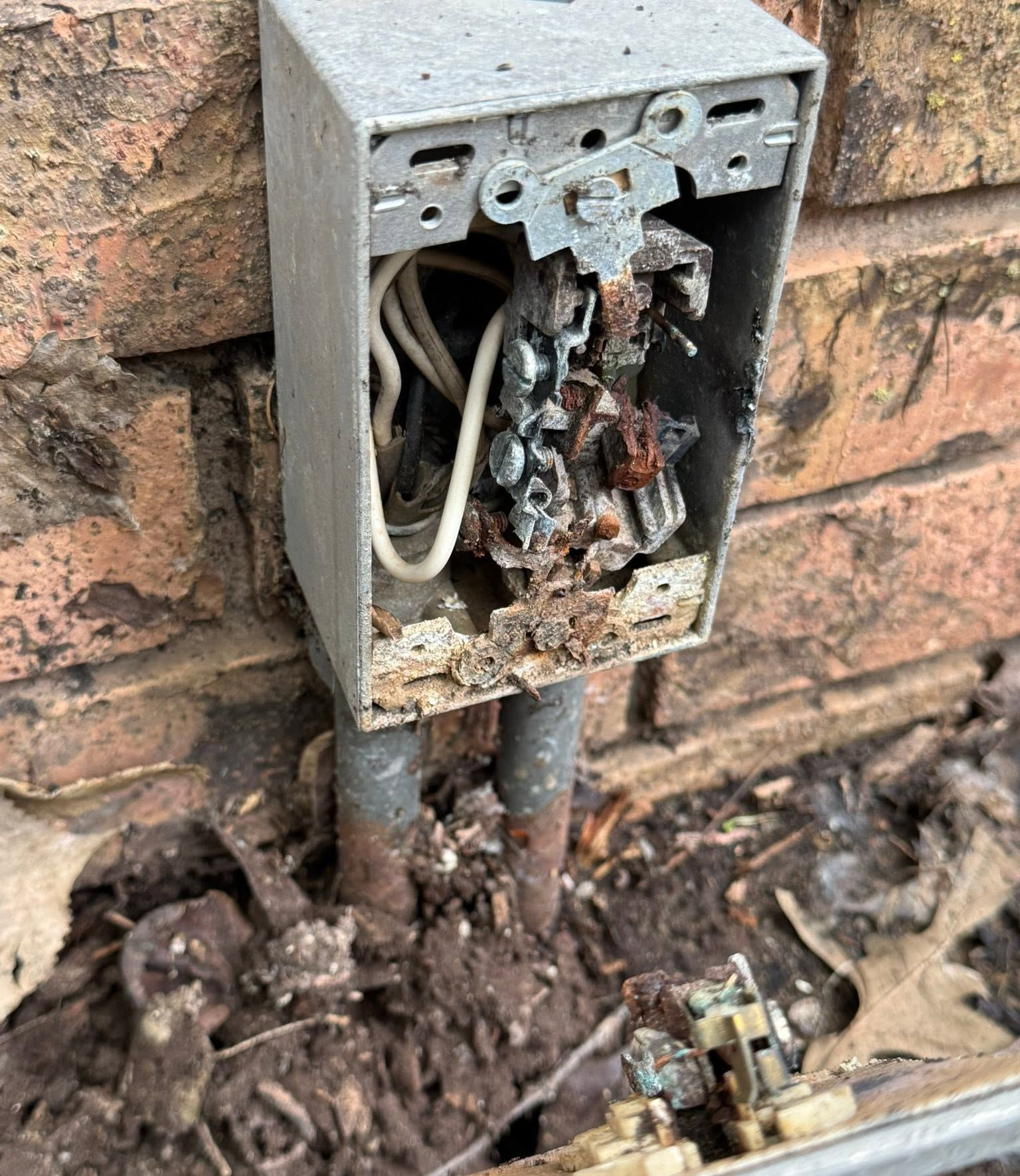 Burned outdoor electrical box mounted on brick wall, with exposed wires and debris below