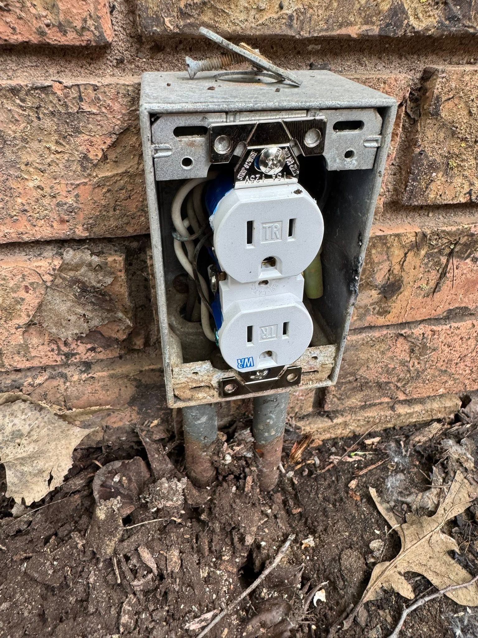 Open electrical outlet box mounted on a brick wall with exposed wiring and dirt below