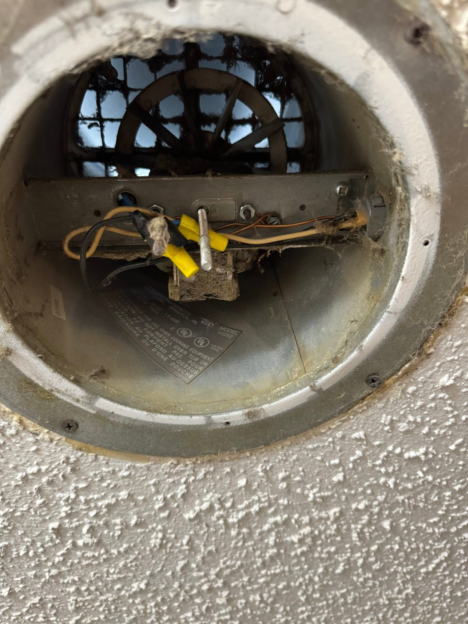 Close-up of a round metal vent opening with exposed wires and yellow tape inside a gray wall