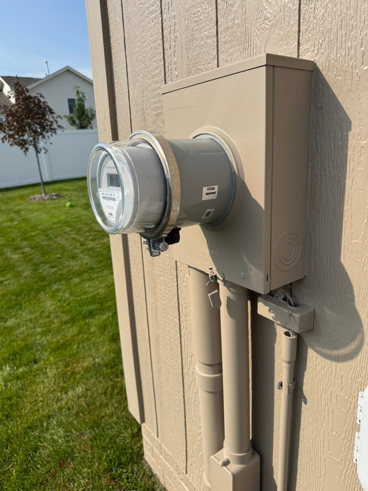 Electric meter on beige house wall beside utility pipes and green lawn