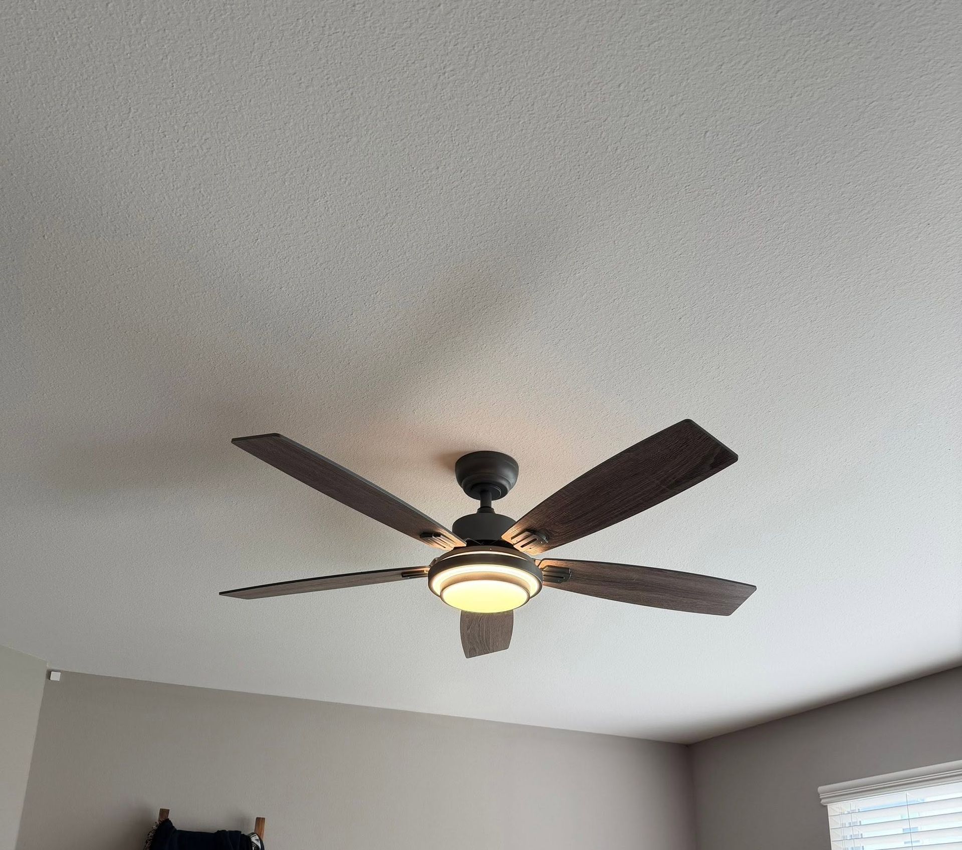 Ceiling fan with dark blades and a lit light fixture in a room with gray walls and a window nearby