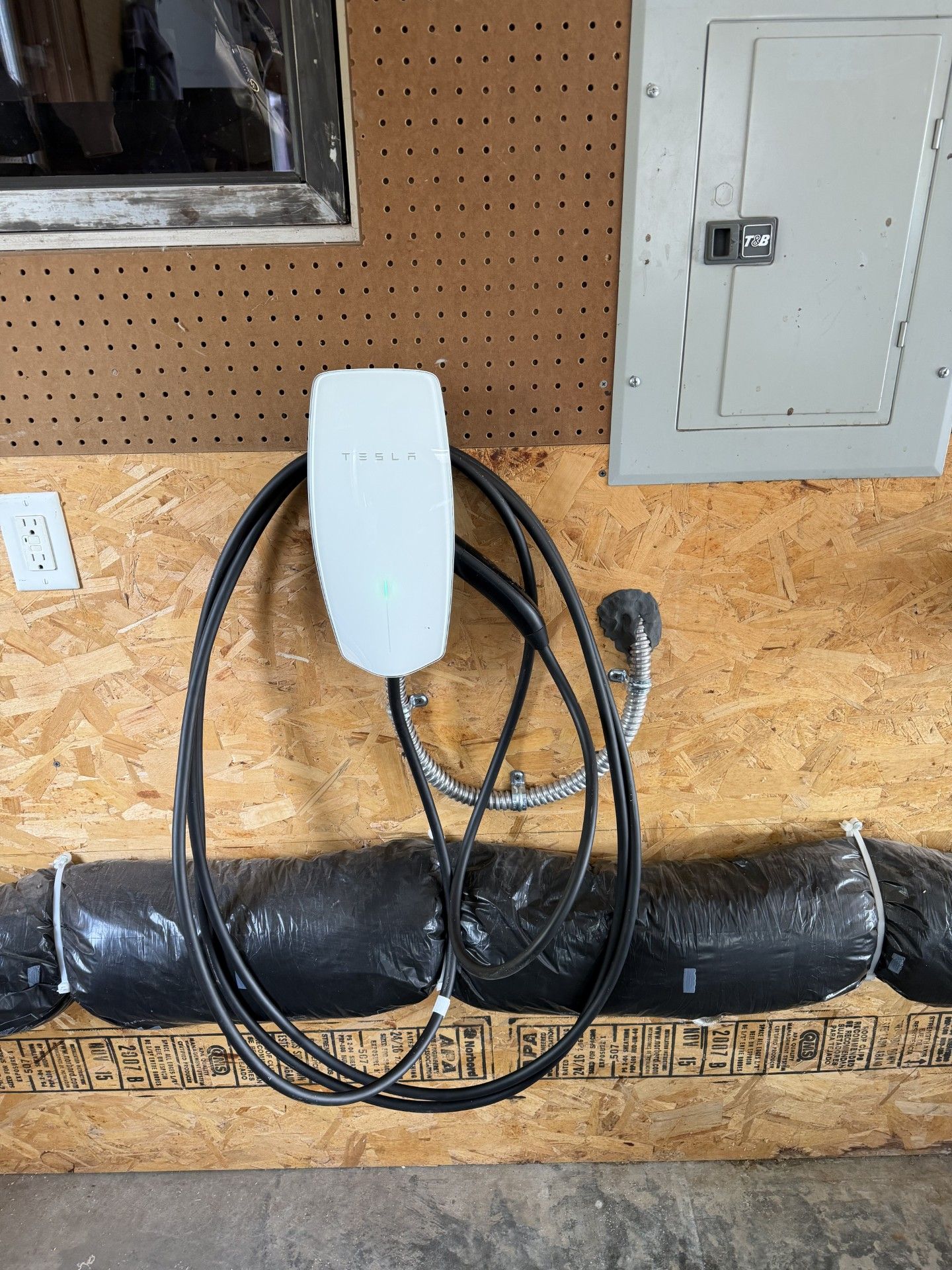 White wall-mounted device with coiled black cables hanging above a black wrapped pipe in a workshop.