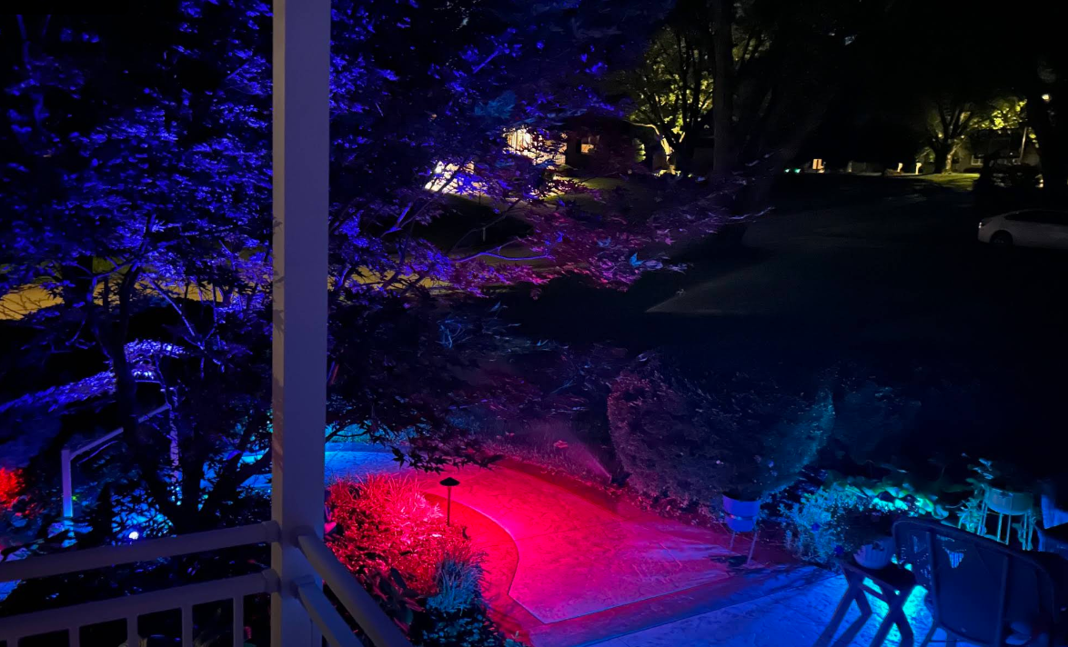 Nighttime garden lit with colorful purple, blue, and red lights around a porch and walkway