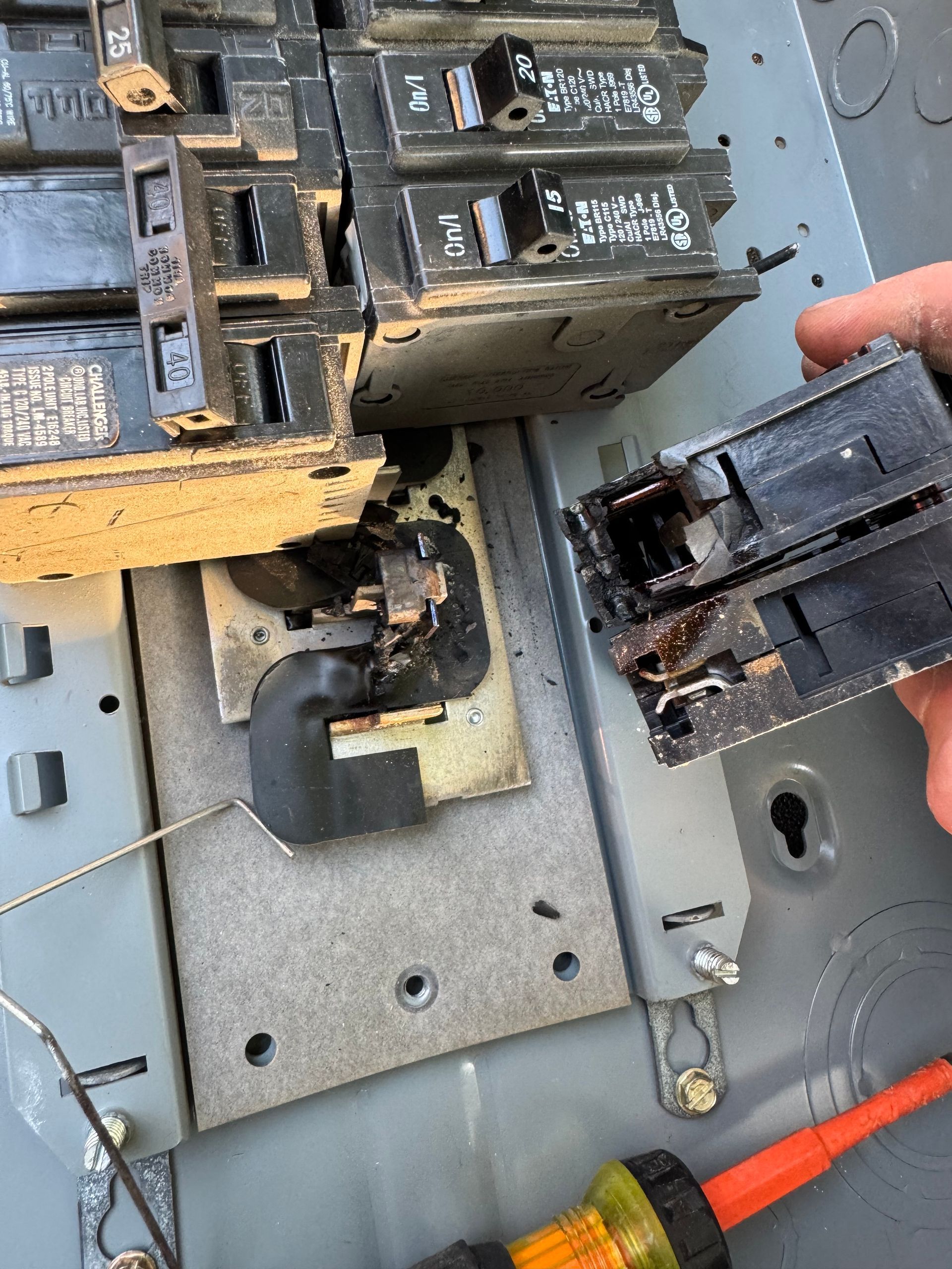 Damaged electrical breaker panel with a burnt breaker and exposed wiring, one breaker held by a hand