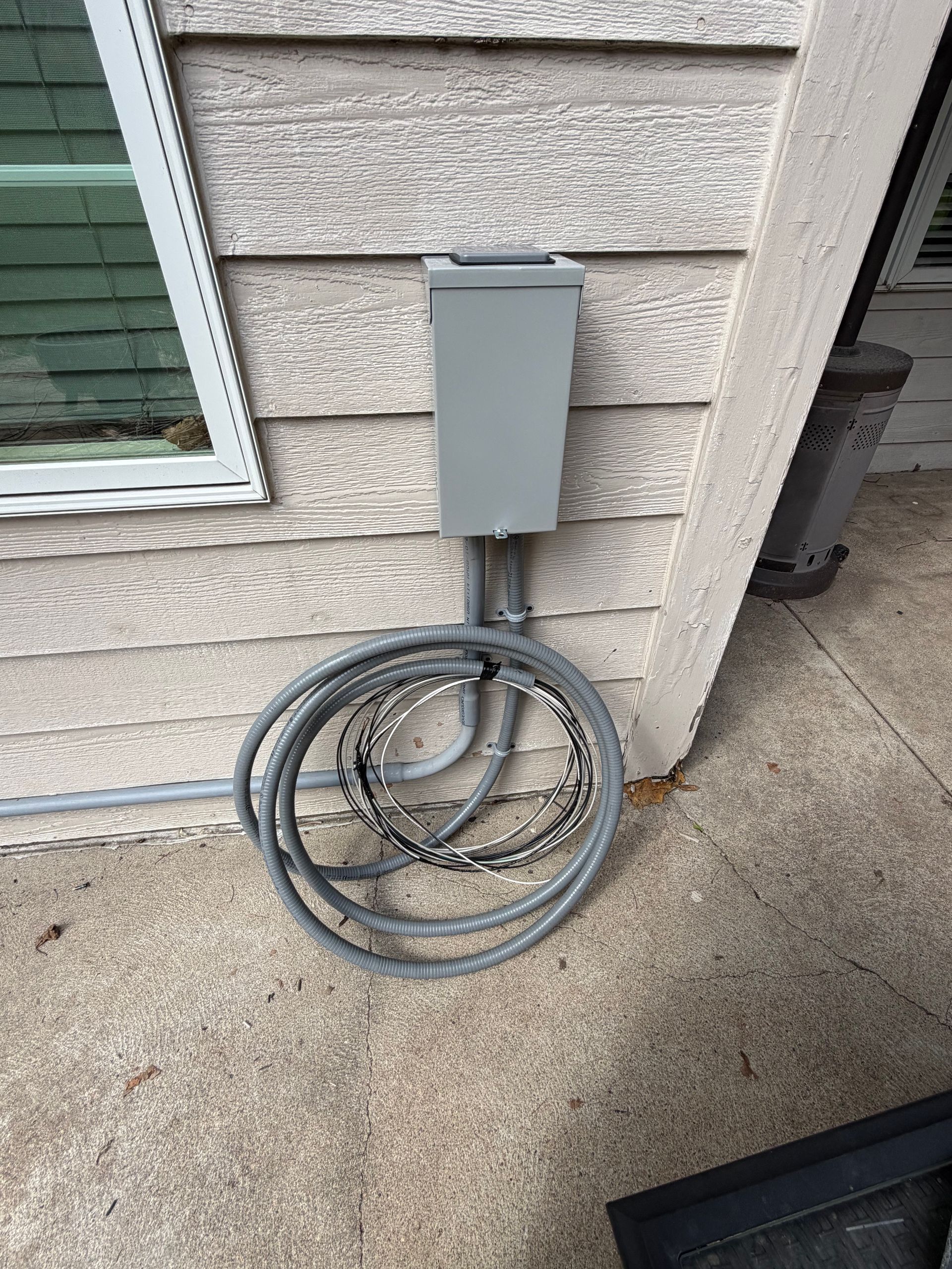 Gray electrical box with coiled cable mounted on an exterior house wall near a window
