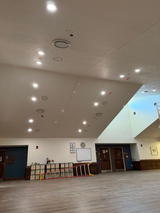 Empty hall with wood floor, recessed ceiling lights, tables and chairs along the back wall, and doors at the right.