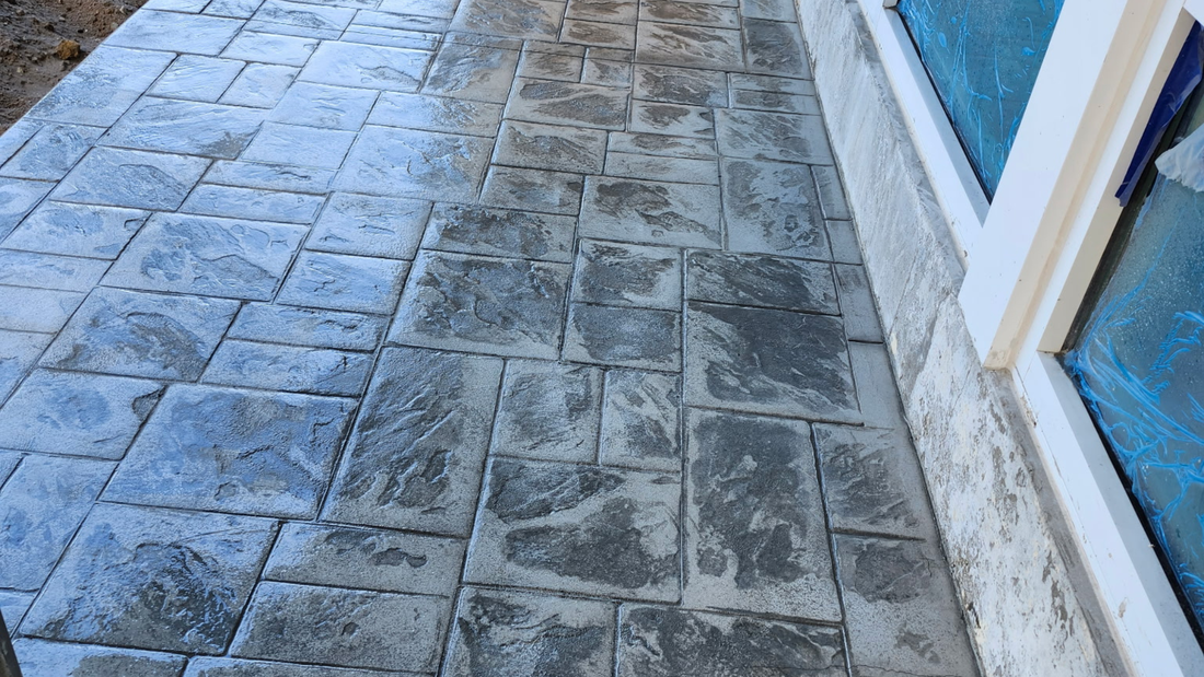 Stamped concrete walkway in gray, adjacent to a building with a white window frame.