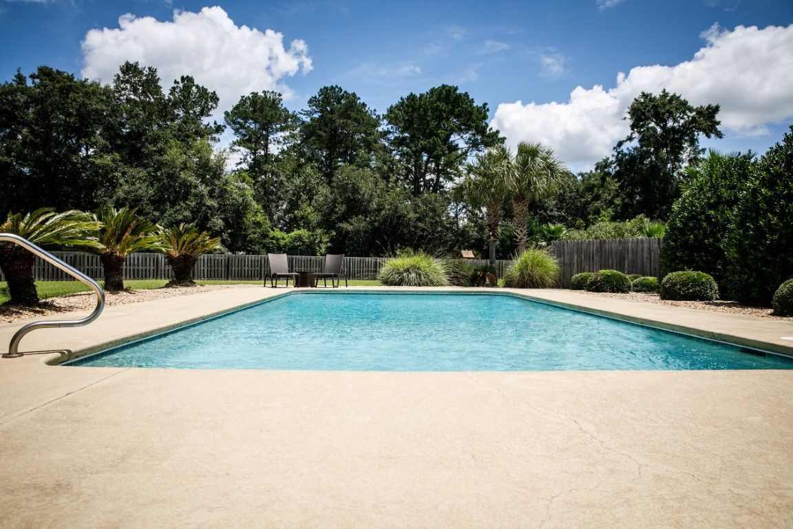 Pool with blue water and surrounding concrete patio, trees, and blue sky with clouds.