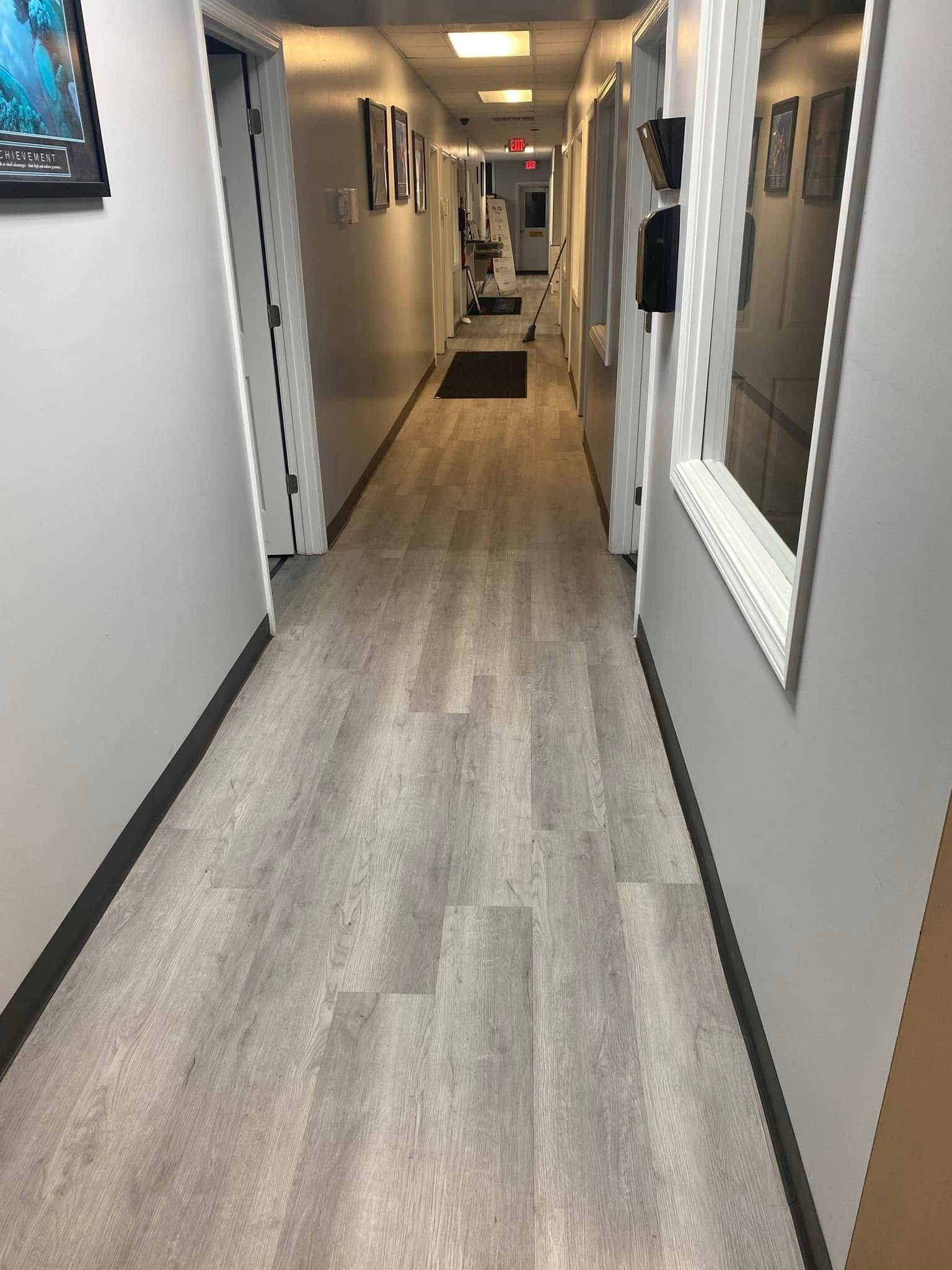 Long hallway with wood-look flooring, gray walls, and framed photos leading to a lit room.