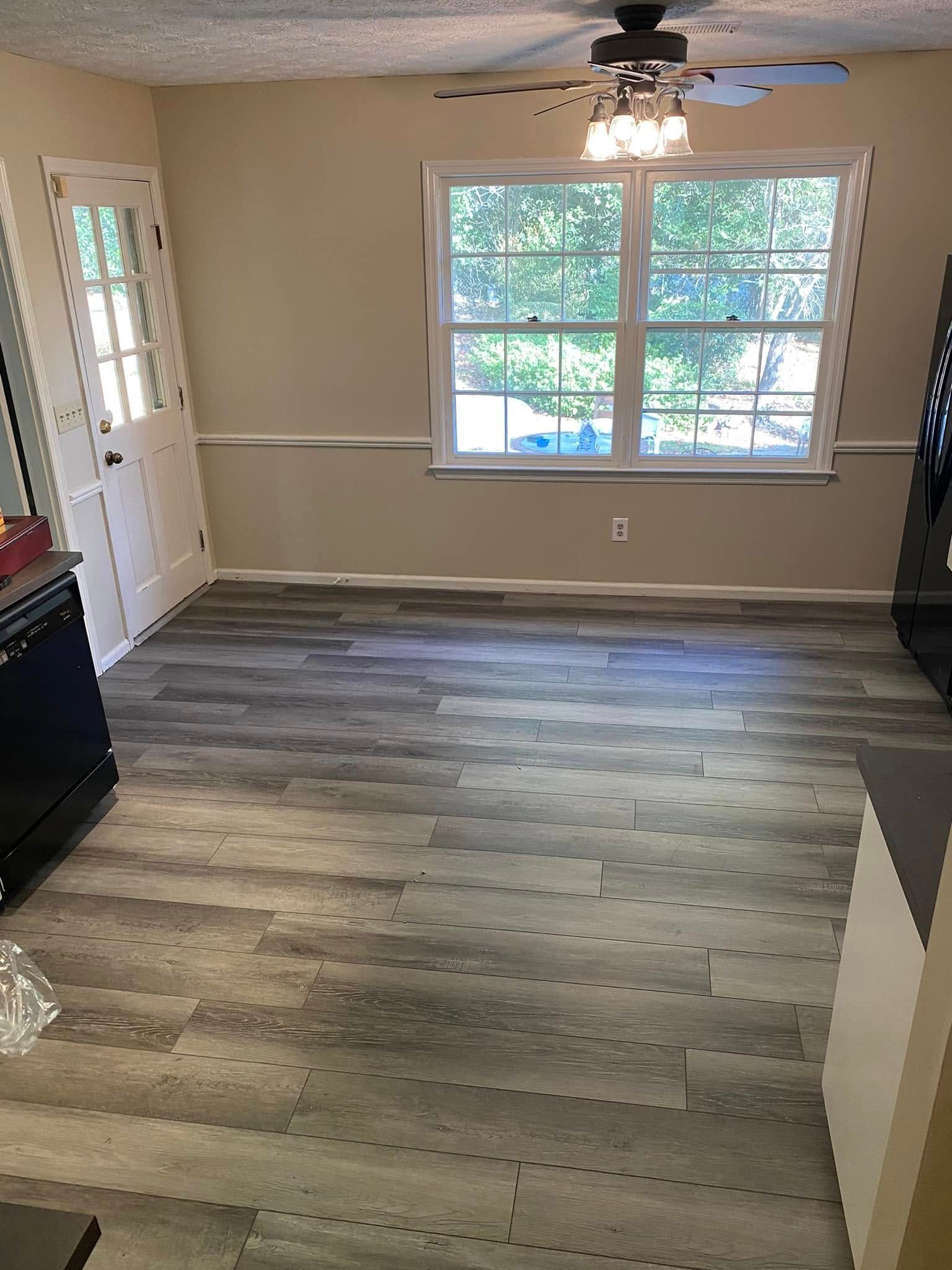 Empty living room with gray wood floors, beige walls, and large window with ceiling fan.