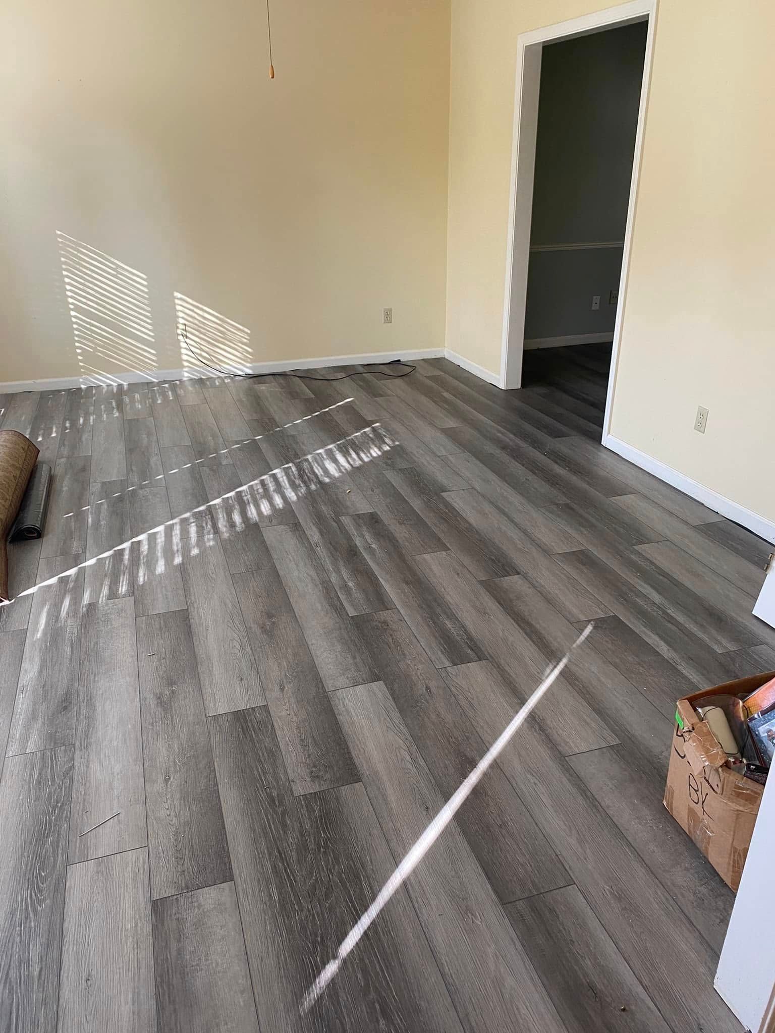 Empty beige room with gray wood flooring and sunlight streaming through blinds onto the floor.