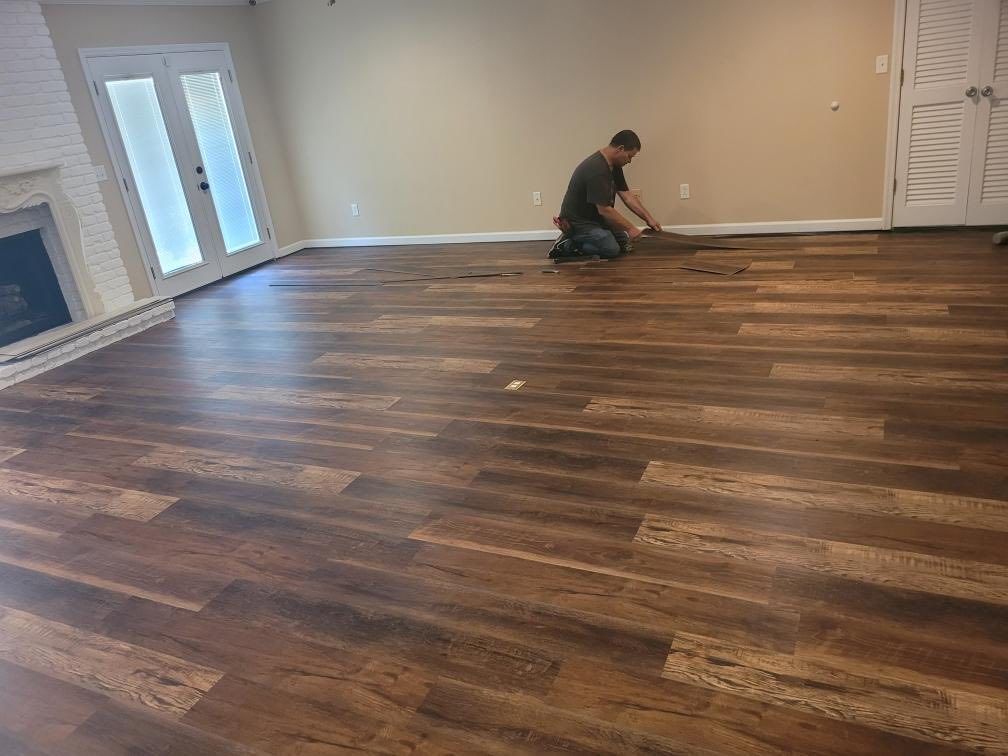 Person installing hardwood flooring in a large empty room with beige walls and white trim.