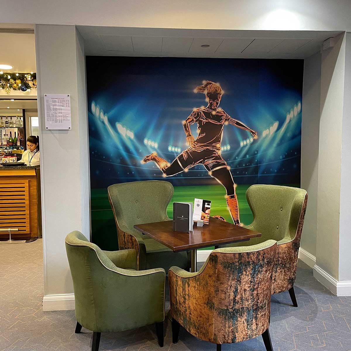 A cafe seating area with a soccer-themed backdrop. Four green armchairs surround a square table.