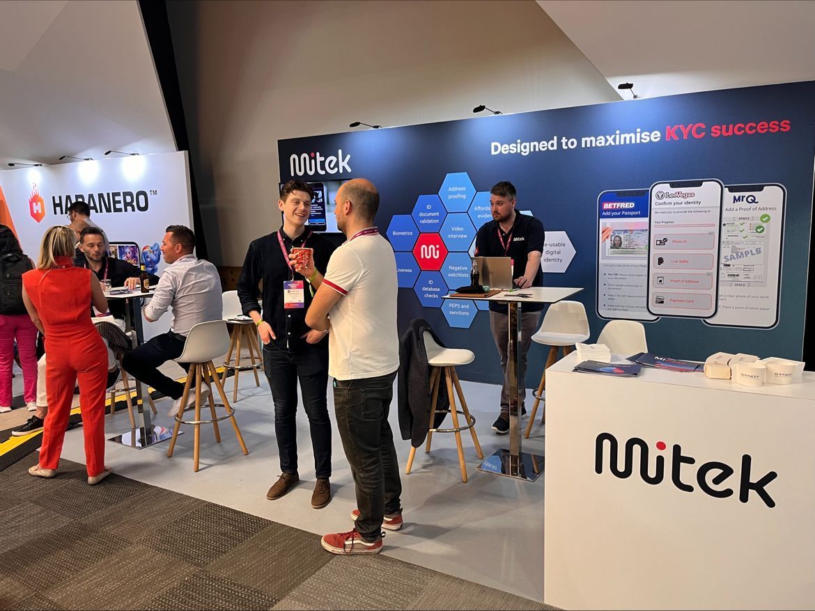 People at the Mitek booth, discussing in a conference setting. Signage and logos are visible.