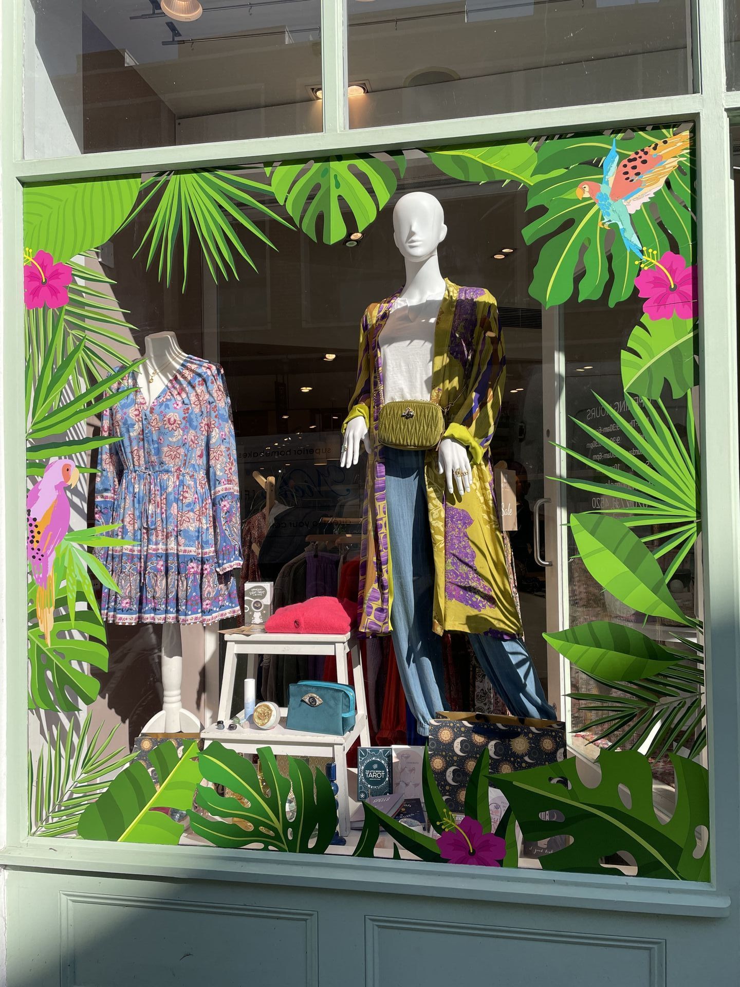 Shop window display: mannequins in tropical-themed outfits surrounded by paper cutouts of leaves and flowers.