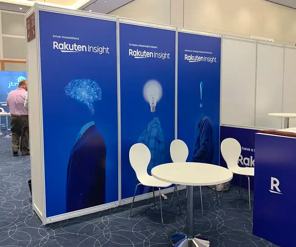Rakuten Insight booth at an event. Blue branding panels, white chairs, and tables are visible.